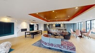 Located at 3575 Silverside Rd in Wilmington, Delaware, this contemporary single-floor amenity space is designed to impress with a bold, upscale aesthetic and a wide range of social and recreational features. The centerpiece of the space is a sprawling open lounge area anchored by a striking tray ceiling finished in a rich burnt orange tone, adding warmth and architectural depth to the room. Light hardwood flooring flows throughout, complementing the bright, airy atmosphere created by an abundance of large, black-framed windows that frame views of the surrounding residential community. Multiple seating arrangements invite gathering and relaxation, including curved sectional sofas, accent chairs, and round metallic coffee tables atop patterned area rugs. A full wet bar with a quartz-topped island, bar stools, stone tile backsplash, and a wall-mounted television serves as a focal point for entertaining. Adjacent to the main lounge, a billiards table with a marble accent wall provides a refined recreational option. A quieter reading or lounge nook with floor-to-ceiling windows and a teal rug offers a more intimate retreat. Sliding panels allow for flexible room configurations, and a separate media or conference room is accessible from the main area. The space balances modern design with inviting comfort throughout.