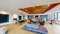 Located at 3575 Silverside Rd in Wilmington, Delaware, this contemporary single-floor amenity space is designed to impress with a bold, upscale aesthetic and a wide range of social and recreational features. The centerpiece of the space is a sprawling open lounge area anchored by a striking tray ceiling finished in a rich burnt orange tone, adding warmth and architectural depth to the room. Light hardwood flooring flows throughout, complementing the bright, airy atmosphere created by an abundance of large, black-framed windows that frame views of the surrounding residential community. Multiple seating arrangements invite gathering and relaxation, including curved sectional sofas, accent chairs, and round metallic coffee tables atop patterned area rugs. A full wet bar with a quartz-topped island, bar stools, stone tile backsplash, and a wall-mounted television serves as a focal point for entertaining. Adjacent to the main lounge, a billiards table with a marble accent wall provides a refined recreational option. A quieter reading or lounge nook with floor-to-ceiling windows and a teal rug offers a more intimate retreat. Sliding panels allow for flexible room configurations, and a separate media or conference room is accessible from the main area. The space balances modern design with inviting comfort throughout.