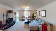 This single-floor residence offers a thoughtfully arranged layout with multiple living spaces connected in an open, flowing configuration. The living room serves as the heart of the home, featuring plush carpeting, a ceiling fan, and generous natural light streaming through large windows that offer views of outdoor greenery. A glass door provides direct access to the exterior. The space comfortably accommodates a large sectional sofa and accent seating, creating a welcoming gathering area. Adjacent to the living room, the dining area features a glass-top table with upholstered seating beneath a multi-light chandelier, with vertical mirror panels along one wall that amplify light and add visual depth. The kitchen is efficiently designed with white cabinetry, tile flooring, a double-basin sink, and an electric range. The primary bedroom offers carpeted flooring, a ceiling fan, vertical blinds, and ample space for a full bedroom suite. A second bedroom functions as a flexible space suitable for sleeping or a home office, also featuring carpet and a ceiling fan. Two full bathrooms are each equipped with a tub-shower combination, vanity with storage, and tile flooring, providing practical functionality throughout the residence.