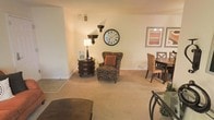 This single-floor residence offers a welcoming and thoughtfully arranged living environment with a classic, timeless aesthetic. The open layout seamlessly connects the living and dining areas, creating a sense of spaciousness ideal for both relaxing and entertaining.

The living area is centered around a plush, patterned wingback armchair accented with a warm terracotta throw pillow, adding a pop of color against the neutral palette. Soft, wall-to-wall carpeting in a warm beige tone runs throughout, contributing to the comfortable and understated ambiance. Decorative wall accents, including a large round clock with Roman numerals and elegant wall-mounted shelving brackets, lend the space a refined, traditional character.

Adjacent to the living area, a dedicated dining space features a wooden table paired with woven rattan-style chairs, offering a cozy spot for meals. Framed artwork on the walls adds visual interest and completes the curated look of the room.

The overall ambiance is warm, calm, and inviting, with consistent neutral tones and classic furnishings that create a cohesive and livable atmosphere throughout the space.