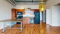 This two-bedroom, two-bathroom unit showcases an industrial-modern aesthetic throughout its single-floor layout. The open-concept living and kitchen area is defined by striking exposed ductwork, a corrugated metal ceiling with track lighting, and warm hardwood flooring that extends throughout the main living space. The kitchen is well-equipped with medium-toned wood cabinetry, granite countertops, black appliances including a range, microwave, dishwasher, and refrigerator, plus a built-in wine rack and a mobile granite-topped island for added functionality.

The main living area is spacious and airy, benefiting from a frosted glass sliding door that adds a contemporary touch while providing flexible room division. Large windows in the bedrooms frame urban rooftop views and flood the rooms with natural light. The bedrooms feature plush carpeting, offering a contrast to the hardwood in the common areas.

Both bathrooms are finished with tile-patterned flooring, rich wood vanity cabinetry, and white countertops. One bathroom includes a tub/shower combo, while the other features a walk-in shower. The overall ambiance is modern and urban, with thoughtful industrial design details lending the space a distinctive loft character.