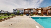 Located at 6205 Longstem Way in Loveland, Colorado, this single-level outdoor community amenity space is thoughtfully designed to encourage relaxation and recreation. At its centerpiece sits a generously sized swimming pool with crystal-clear blue water, complemented by an adjacent spa, offering residents a resort-style experience beneath Colorado's expansive open skies.

Surrounding the pool is a broad concrete deck furnished with lounge chairs and shaded by bold umbrella canopies, providing ample space for sunbathing and socializing. Elegant wood pergola structures with latticed privacy screens define dedicated seating areas, adding warmth and architectural character to the space. Additional casual seating zones with patio furniture and shade umbrellas are distributed throughout the grounds.

Toward one side of the amenity area, a well-maintained bocce ball court offers a dedicated recreational activity zone. The overall layout, visible from the aerial scan, reveals a cohesive and well-organized outdoor environment with mulched landscaping beds, young trees, and defined pathways connecting each feature area. A secure black metal perimeter fence encloses the entire space, ensuring privacy and safety. The backdrop of the surrounding residential neighborhood, with its modern farmhouse-style homes, frames the amenity area and contributes to the welcoming, community-oriented atmosphere.