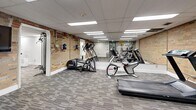 Located at 40 Vanier Dr in Guelph, Ontario, this fitness center offers a thoughtfully designed workout space spread across a single floor. The facility features a drop-panel ceiling with recessed and flush-mounted fluorescent lighting that ensures the space is consistently and evenly lit throughout. Durable, patterned carpet tile flooring covers the entire area, providing comfort underfoot during workouts.

The space is divided into functional zones for various exercise types. A dedicated cardio area houses multiple treadmills, elliptical trainers, and a stationary bike, complemented by full-length wall mirrors that enhance the sense of openness. An exposed brick wall runs along one side, adding a warm, industrial character that contrasts with the otherwise clean, modern white walls.

The free weights section is well-stocked with a full dumbbell rack, an adjustable workout bench, and a wall-mounted mirror for form monitoring. A motivational typographic mural featuring words such as "Dedication," "Commitment," and "Push" adds an energizing visual element. A separate area stores stability balls and additional accessories, with two private rooms accessible via glass-panel doors — suitable for stretching, personal training, or storage. Another vivid mural reading "Live Well, Live Strong" anchors the cardio zone, reinforcing the facility's active and motivating atmosphere.