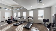 Located at 1340 Farnham Point in Colorado Springs, CO, this fitness center offers a comprehensive and thoughtfully designed space for a full-body workout experience. The single-level facility is bathed in natural light through rows of large windows fitted with white blinds, framing stunning views of the surrounding Colorado mountains and providing an energizing atmosphere throughout the day.

The main workout area features durable patterned carpet flooring and neutral gray walls that lend a clean, modern aesthetic. Exposed industrial-style HVAC ductwork runs along the upper walls, adding a contemporary architectural element beneath the recessed-lit ceiling. The space is generously equipped with multiple treadmills, elliptical trainers, and stationary bikes positioned to take advantage of the scenic window views.

A dedicated strength training zone houses a multi-station cable machine, a leg press, a flat workout bench, and a well-stocked dumbbell rack with a full range of weights. A floor mat area provides space for stretching and bodyweight exercises. Near the entry, a glass-paneled door and interior window connect the fitness center to an adjacent common area, offering visual openness. A water fountain is conveniently positioned near the entrance. An exterior door provides direct outdoor access, completing this well-appointed and functional community fitness facility.