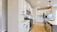 This two-story home presents a clean, contemporary design carried consistently throughout its spaces. The main level is anchored by an open-concept kitchen and living area, where light hardwood flooring extends seamlessly underfoot. The kitchen is a standout feature, outfitted with white shaker-style cabinetry accented by matte black hardware, a classic white subway tile backsplash, and light quartz countertops. A central island finished in a soft blue-grey tone provides additional workspace and seating, illuminated overhead by stylish pendant lights. Stainless steel appliances, including a gas range, over-range microwave, and French door refrigerator, complete the functional kitchen setup. A fireplace anchors the living area, adding warmth to the open layout. A craftsman-style entry door with a small window pane welcomes natural light into the entryway. Upper-level bedrooms feature neutral carpet, warm-toned walls, and windows fitted with white blinds that allow ample natural light. Bathrooms on both levels are finished with light hardwood-style flooring, white vanities with quartz tops, and tub-shower combinations. The lower level includes an unfinished basement with concrete walls and floors, offering significant potential for customization.