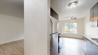 Located in Portland, OR, this single-floor, 2-bedroom, 1-bathroom unit offers a practical and well-organized layout. The combined kitchen and living area forms the heart of the home, featuring light wood-style flooring throughout. The kitchen is equipped with dark cabinetry and ample storage in one configuration, while another variation showcases sleek grey cabinetry with white countertops, a stainless steel range, and a ceiling fan overhead. A large window and sliding glass door flood the living area with natural light and provide direct access to a private balcony overlooking lush greenery. The spacious primary bedroom features plush grey carpeting, a wide window with outdoor views, and a baseboard heater. The second bedroom also features carpet and a sliding-door closet, offering practical storage. A connecting hallway links the bedrooms to the shared full bathroom, which includes a vanity with dark wood cabinetry, wood-style tile flooring, and grab bars for accessibility. Throughout the unit, clean white walls and neutral tones create a bright, cohesive atmosphere well-suited for comfortable everyday living.