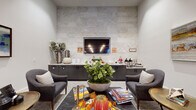 This single-floor community space is designed to foster comfort and social connection through a refined, contemporary aesthetic. The lounge area serves as an inviting focal point, featuring plush upholstered armchairs arranged around a sleek coffee table atop a patterned area rug. Wood-clad columns add warmth and architectural character, while curated artwork—including abstract landscape paintings in rich blues and earthy tones—adorns the neutral walls, contributing to a calm and welcoming ambiance.

A standout feature of the lounge is the full-wall accent panel finished in textured stone tile with a geometric relief pattern. Set within this striking backdrop is a wall-mounted flat-screen television above a long granite-topped service counter with dark cabinetry. This refreshment station is stocked with coffee dispensers, snacks, and seasonal décor, offering a casual gathering spot for residents or guests.

The adjacent dining area is expansive and airy, with high ceilings dressed in a moody, cloud-patterned finish and illuminated by a series of elegant pendant lights. Round and rectangular tables paired with neutral and yellow upholstered chairs fill the space, which benefits from natural light streaming through large exterior-facing windows and glass doors. Herringbone-patterned tile and plush carpet define separate zones, lending the overall space a polished yet approachable character.