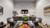 This single-floor community space is designed to foster comfort and social connection through a refined, contemporary aesthetic. The lounge area serves as an inviting focal point, featuring plush upholstered armchairs arranged around a sleek coffee table atop a patterned area rug. Wood-clad columns add warmth and architectural character, while curated artwork—including abstract landscape paintings in rich blues and earthy tones—adorns the neutral walls, contributing to a calm and welcoming ambiance.

A standout feature of the lounge is the full-wall accent panel finished in textured stone tile with a geometric relief pattern. Set within this striking backdrop is a wall-mounted flat-screen television above a long granite-topped service counter with dark cabinetry. This refreshment station is stocked with coffee dispensers, snacks, and seasonal décor, offering a casual gathering spot for residents or guests.

The adjacent dining area is expansive and airy, with high ceilings dressed in a moody, cloud-patterned finish and illuminated by a series of elegant pendant lights. Round and rectangular tables paired with neutral and yellow upholstered chairs fill the space, which benefits from natural light streaming through large exterior-facing windows and glass doors. Herringbone-patterned tile and plush carpet define separate zones, lending the overall space a polished yet approachable character.