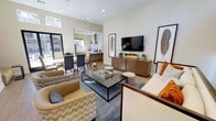 This single-floor space showcases a thoughtfully designed open-concept layout that seamlessly connects the living, dining, and kitchen areas into one cohesive and inviting environment. The living area is anchored by a sleek media console in a warm wood finish, paired with a wall-mounted flat-screen television. Comfortable seating, including plush armchairs in tan and patterned upholstery, surrounds a minimalist rectangular coffee table set atop a blue geometric area rug, creating a relaxed yet refined gathering space. Accent pillows in burnt orange add a pop of color against the neutral palette of cream and beige tones that define the interior.

The dining area flows naturally from the living space and features a wood dining table with upholstered chairs, positioned near large windows that invite abundant natural light and offer views of lush greenery outside. Pendant lighting above the dining zone adds an elegant touch.

The kitchen, visible in the background, is outfitted with modern cabinetry in a warm wood tone, a statement range hood, and ample counter space, reinforcing the home's contemporary aesthetic. High ceilings amplify the sense of openness, while recessed lighting throughout ensures a bright and welcoming ambiance. Light hardwood flooring ties the entire space together with warmth and continuity.