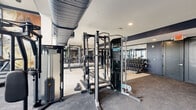 This fitness center offers a comprehensive, single-level workout facility with a generously sized open floor plan designed to accommodate a wide variety of exercise disciplines. The space is defined by soaring double-height ceilings along the cardio zone, where floor-to-ceiling windows flood the interior with abundant natural light and provide outdoor views, creating an energizing atmosphere for training.

The cardio section lines the windowed wall and houses a row of elliptical trainers and stationary bikes. A large-scale abstract mural serves as a bold focal point in this area, adding a dynamic artistic element to the environment. Exposed industrial-style ductwork runs overhead, contributing to the space's contemporary aesthetic alongside the navy blue accent walls and neutral grey carpeting throughout.

The strength training zone is well-appointed with Precor multi-functional cable machines, a smith machine, barbell racks with weight plates, and multiple dumbbell racks ranging across a broad weight selection. Large wall mirrors enhance the sense of depth within the strength area.

A dedicated support station along the navy-painted rear wall includes built-in open shelving, a mini-refrigerator, and a wall-mounted television, providing practical amenities for users. The overall design balances function and style effectively.