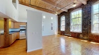 This two-story, 3-bedroom, 2-bathroom apartment showcases a compelling blend of historic industrial character and functional modern living. The expansive main living area immediately impresses with soaring ceilings, exposed wooden beam ceilings with tongue-and-groove planking, and striking original brick walls punctuated by tall arched and multi-pane windows that flood the space with abundant natural light. Warm hardwood floors run throughout the main level, complementing the raw, loft-style aesthetic. A ceiling fan adds a practical touch to the voluminous living space.

The kitchen features a U-shaped layout with ample light maple cabinetry, generous granite-style countertops, and a full suite of appliances, including a range with overhead ventilation. A pass-through counter connects the kitchen to the open living area, maintaining the sense of openness.

The upper level, accessed via a staircase with a wood handrail, offers carpeted bedroom space beneath the same distinctive wood-planked ceiling, with exposed brick visible from the upper landing. Two full bathrooms are present, each featuring a tub/shower combination — one with rich hardwood flooring and a pedestal sink, the other with tile flooring. Together, these elements create a distinctive, character-rich living environment.