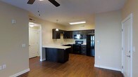 This single-floor residence offers a well-designed open-concept layout that seamlessly connects the kitchen and living areas into one cohesive space. The kitchen is a standout feature, showcasing rich dark espresso cabinetry that extends to the upper walls, providing ample storage alongside a clean, light-colored countertop that creates a striking contrast. A peninsula breakfast bar extends from the cabinetry, offering additional prep space and a casual seating area. The kitchen is fully equipped with a suite of black appliances, including a refrigerator, electric range, built-in microwave, and a functional sink positioned beneath a pendant light that adds a warm, focused glow to the workspace. A skylight-style ceiling fixture brightens the kitchen naturally from above. Warm hardwood-style flooring flows continuously throughout the space, adding depth and warmth to the overall aesthetic. The living area is complemented by a ceiling fan, ensuring comfort year-round. A hallway leads to additional rooms, including an arched-panel door that adds a classic architectural touch. Neutral warm-toned walls keep the space feeling bright, open, and inviting throughout.