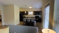 This single-floor space is designed with an open-concept layout that seamlessly connects the living and kitchen areas, creating a cohesive and functional environment. The kitchen is a standout feature, showcasing rich dark espresso cabinetry that contrasts beautifully against the light-toned walls. It is fully equipped with black appliances, including a refrigerator, microwave, and range, alongside dark countertops that add a sleek, modern touch. A central kitchen island serves as the focal point, offering additional counter space and casual seating with dark wood bar-height chairs — ideal for dining or entertaining. Pendant lighting above the island and a flush-mount ceiling fixture provide warm, layered illumination throughout the kitchen. The living area, visible from the kitchen, features a comfortable seating arrangement and benefits from natural light streaming in through slatted blinds on a large window. Light wood-tone flooring runs throughout, tying the spaces together and enhancing the open, airy feel of the interior. The overall ambiance is modern and welcoming, with a neutral color palette and thoughtful design details that balance practicality with everyday comfort.
