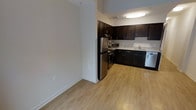 This single-floor space is designed with both functionality and style in mind. The kitchen is a standout feature, showcasing rich dark espresso-toned cabinetry that extends from base to upper wall units, providing generous storage throughout. Light-toned countertops offer a striking contrast against the dark cabinetry, while a full suite of stainless steel appliances — including a refrigerator, range, microwave, and dishwasher — lends a modern, polished look to the cooking area. The kitchen is arranged in an efficient L-shaped layout, maximizing workspace while maintaining an open feel. Flush-mounted ceiling lighting illuminates the kitchen area effectively, creating a bright and welcoming environment. Light hardwood-style flooring flows seamlessly throughout the space, adding warmth and visual continuity between the kitchen and the adjacent open area. The neutral, light-toned walls complement the flooring and contribute to a clean, airy ambiance. The open transitional area adjacent to the kitchen provides flexible space that can accommodate a variety of living arrangements. Overall, the space offers a practical yet refined interior with a cohesive design aesthetic.