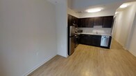 This single-floor space is designed with both functionality and style in mind. The kitchen is a standout feature, showcasing rich dark espresso-toned cabinetry that extends from base to upper wall units, providing generous storage throughout. Light-toned countertops offer a striking contrast against the dark cabinetry, while a full suite of stainless steel appliances — including a refrigerator, range, microwave, and dishwasher — lends a modern, polished look to the cooking area. The kitchen is arranged in an efficient L-shaped layout, maximizing workspace while maintaining an open feel. Flush-mounted ceiling lighting illuminates the kitchen area effectively, creating a bright and welcoming environment. Light hardwood-style flooring flows seamlessly throughout the space, adding warmth and visual continuity between the kitchen and the adjacent open area. The neutral, light-toned walls complement the flooring and contribute to a clean, airy ambiance. The open transitional area adjacent to the kitchen provides flexible space that can accommodate a variety of living arrangements. Overall, the space offers a practical yet refined interior with a cohesive design aesthetic.