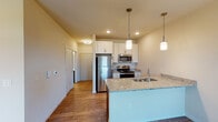 This single-floor unit offers a thoughtfully designed open-concept layout that seamlessly connects the kitchen and living areas. The kitchen is a standout feature, equipped with stainless steel appliances including a refrigerator, range, and microwave. White upper cabinetry contrasts elegantly with rich dark lower cabinets, while light granite countertops extend across both the prep surfaces and a peninsula island. Pendant lighting hangs above the island, adding a warm, focused glow to the space. Hardwood-style flooring runs throughout the kitchen and adjoining hallway, lending a clean, cohesive look.

The living area transitions to plush carpeting, creating a comfortable and inviting atmosphere. A sliding glass door opens to a private balcony, bringing in natural light and offering views of surrounding greenery. A side window further enhances the brightness of the room.

A built-in closet with open shelving provides convenient storage directly off the living space. The bathroom features light wood-style flooring, a dark wood vanity with a white countertop and sink, and a clean, elongated layout that maximizes functionality. Neutral wall tones throughout the unit create a calm and versatile backdrop, making the space feel open and move-in ready.
