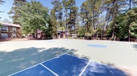 This outdoor recreational space features a well-maintained, dual-purpose sport court designed to accommodate both basketball and tennis. The court surface is finished in a combination of blue and green, with clearly marked boundary lines for each sport, ensuring organized and versatile use. A full basketball key and arc are visible toward the center and rear of the court, while tennis court lines are laid out across the foreground, maximizing the utility of the space.

The court is surrounded by a generous canopy of mature trees, including tall pines and leafy deciduous varieties, which provide natural shade and a serene, park-like setting. The lush greenery creates a natural boundary around the perimeter, offering a sense of privacy and enclosure while maintaining an open, airy feel under a clear blue sky.

In the background, multi-story residential buildings are visible through the trees, suggesting this court serves as a shared amenity within a larger residential community. A low perimeter fence lines the far edge of the court, separating it from the surrounding landscaping. The overall ambiance is vibrant and inviting, offering residents an accessible and well-equipped outdoor space for active recreation in a naturally scenic environment.
