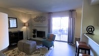 This single-floor residence offers a thoughtfully arranged living area that balances comfort and functionality. The main living room serves as the centerpiece of the space, highlighted by a classic white-mantled fireplace that adds warmth and character to the room. Hardwood-style flooring runs throughout, providing a clean and cohesive aesthetic underfoot.

Natural light flows generously into the living area through large sliding glass doors, which are framed by decorative patterned curtains. The doors provide direct access to an outdoor patio, where seating is visible, extending the livable space outdoors and offering pleasant views of the surrounding greenery.

The living room is finished with a textured ceiling and neutral-toned walls accented by decorative wall art, creating an inviting ambiance. A floor lamp in the corner adds layered lighting options for the space. The layout accommodates a comfortable seating arrangement, with room for an area rug that anchors the space visually.

Overall, this residence presents a warm and functional single-level layout that emphasizes natural light, classic architectural details such as the fireplace mantel, and a seamless connection between indoor living and outdoor spaces.