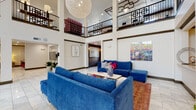 Located at 6600 McKinney Ranch Pkwy in McKinney, Texas, this two-story community clubhouse is designed to impress from the moment you enter. The grand lobby commands attention with soaring double-height ceilings, large globe pendant light fixtures, and intricate geometric ceiling medallions. Rich, white tile flooring anchors the open seating area, furnished with bold cobalt-blue sectional sofas atop a patterned area rug, complemented by vibrant artwork — including a striking equestrian canvas — throughout. Floor-to-ceiling glass doors at the rear of the lobby flood the space with natural light and offer views of the surrounding grounds. A branded elevator and staircase provide seamless access between floors. The leasing area features warm wood-toned workstations with a sheer decorative partition, dark hardwood flooring, and ornate lattice ceiling detailing carried throughout the upper level. The fully equipped kitchen showcases an expansive marble-topped island with bar seating, pendant lighting, taupe cabinetry, stainless appliances, and rich hardwood floors. A cozy lounge nook houses dual flat-screen TVs with playful circuit-tree wall art. The second level opens to a dedicated recreation corridor complete with a shuffleboard table, arcade games, and a casual bistro seating area, all overlooking the double-height lobby below through elegant wrought-iron railings.