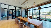 Located at 150 River St in Hackensack, NJ, this contemporary single-floor space is designed with a sophisticated blend of modern aesthetics and functional versatility. The interior is anchored by an expansive co-working area featuring a long, warm-toned communal table surrounded by distinctive plaid-upholstered barrel stools, all illuminated by an eye-catching arrangement of mixed pendant lights with glass and metallic shades. Floor-to-ceiling windows span the perimeter, flooding the space with natural light and offering open views of the surrounding landscape. A lounge zone with leather and neutral-toned seating provides a comfortable retreat alongside the work areas. A separate dining or bar section showcases sleek black high-top tables and padded counter chairs set against large windows, accented by sculptural ring pendant fixtures and exposed brick walls. A moodier corridor area features rich, textured dark walls, wood-look tile flooring, and curated artwork, lending an intimate contrast to the brighter main hall. The grand entry features soaring ceilings, full-height glass doors, and a sculptural driftwood accent piece, creating a striking first impression. Light hardwood flooring flows throughout the primary areas, unifying the space with warmth and continuity. Overall, this space balances professional functionality with upscale, design-forward interiors.