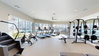 Located at 3755 E McKinney St in Denton, TX, this fitness center offers a comprehensive, single-floor workout facility designed to support a wide range of exercise routines. The space is bright and open, with large floor-to-ceiling windows lining one side of the room, flooding the interior with natural light and providing views of the outdoor pool area beyond.

The cardio zone features multiple treadmills, stationary bikes, a stair climber, and an elliptical, all positioned to take advantage of the scenic window views. The strength training area is equally well-appointed, with a multi-station cable machine, a Smith machine/power rack with weight plates, a full dumbbell rack ranging across multiple weight increments, and adjustable workout benches. Full-length mirrors on one wall enhance the sense of space and support proper form during training.

The interior walls feature a bold, colorful geometric mural accent in teal, yellow, and red, adding an energetic visual element to the otherwise neutral color palette of gray and white. Durable speckled flooring throughout provides a practical and clean surface suited for high-traffic use. Ceiling fans with recessed lighting ensure comfortable air circulation and consistent illumination. The layout is spacious and well-organized, accommodating both cardio and strength training needs efficiently within the footprint.