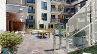 Located at 3170 W Olympic Blvd in Los Angeles, this contemporary amenity space is designed to foster community and comfort within a modern multi-story residential building. At its heart lies a spacious open-air courtyard, enclosed by the building's striking facade of clean white panels, warm wood accents, and steel-framed balconies. The courtyard is thoughtfully appointed with plush outdoor seating arrangements, a wooden dining table with seating, and raised planter beds filled with vibrant ornamental grasses and tropical plants, all set on large-format stone tile flooring. Glass stair railings add a refined architectural touch throughout the outdoor area.

Seamlessly accessible from the courtyard, the indoor lounge area features a warm wood-paneled ceiling, a sleek linear fireplace set within a built-in light wood cabinetry wall, and a large mounted flat-screen television. Comfortable gray armchairs and bold mustard ottomans provide an inviting, curated atmosphere.

An adjacent kitchen and gathering space boasts a generous island with bar seating, floor-to-ceiling cabinetry, stainless steel appliances, and a striking wood-slat ceiling that extends to large sliding glass doors opening directly to the courtyard. The space also includes a clean, spa-inspired shower room finished with white subway tile walls and large-format stone flooring.