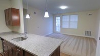 This single-floor space showcases an open-concept design that seamlessly connects the kitchen and living areas. The kitchen features a granite-topped breakfast bar with dark cabinetry and hardwood-style flooring, illuminated by two elegant pendant lights hanging overhead. A flush-mount ceiling fixture provides additional ambient lighting. The living area transitions to plush carpeting and is brightened by a glazed exterior door and a large window fitted with blinds, allowing natural light to fill the space. The neutral, light-toned walls throughout create a clean and welcoming atmosphere. The layout offers a practical flow between the kitchen and living zone, making the space feel open and functional. Visible HVAC vents in the ceiling indicate a climate-controlled environment. Overall, the space presents a well-maintained, move-in-ready interior with a balance of modern finishes and comfortable living features.
