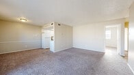 This single-floor residential unit offers a clean, functional layout designed for comfortable everyday living. The open living area is generously sized, featuring neutral-toned carpet that extends throughout most of the space, warm beige walls, and a textured ceiling. Natural light enters through a sliding glass door fitted with vertical blinds, which opens to an outdoor patio area, as well as through an additional window that brightens the room further. The living area flows seamlessly from the kitchen, which is positioned along one side and features a galley-style configuration with white cabinetry, brown laminate countertops, a stainless steel sink, a white range, and wood-look vinyl flooring that provides a clean contrast to the carpeted areas. The bedroom is a well-proportioned room with plush carpet, a ceiling fan, and its own natural light source via a window with blinds. It includes direct access to both a private bathroom and a walk-in closet, offering practical storage and added convenience. The bathroom is accessible through a dedicated doorway from the bedroom. Throughout the unit, neutral tones and simple finishes create a cohesive, understated aesthetic. Climate control is supported by wall-mounted thermostats and ceiling vents visible in multiple rooms.
