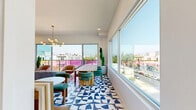 Located at 360 N Hayworth Ave in the heart of Los Angeles, this single-floor amenity space delivers a bold, contemporary aesthetic that blends lively color with modern design sensibility. The standout feature throughout is the striking geometric blue-and-white patterned tile flooring, which anchors the entire space and sets a vibrant visual tone.

The central dining area comfortably accommodates a group around a natural wood table paired with teal upholstered chairs, all bathed in abundant natural light streaming through large sliding glass doors and expansive floor-to-ceiling windows. These openings frame sweeping views of the Los Angeles skyline and surrounding neighborhoods, creating an airy, open atmosphere. A sculptural brass and globe pendant chandelier adds a warm, sophisticated touch overhead.

A lounge area complements the dining space, furnished with a plush blue sofa, rounded accent chairs in blush and caramel tones, and a built-in banquette with tan leather panels. A compact kitchenette features deep navy cabinetry, marble-look surfaces, an under-counter beverage cooler, and a matte black faucet — offering functional convenience with refined style.

The bathroom is clean and minimal, finished in white tile with large-format flooring, a wall-mounted sink, black-framed mirror, and sleek vertical sconce lighting. A connecting hallway provides smooth flow between all areas of the space.
