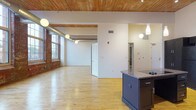 This studio loft showcases a thoughtful blend of industrial heritage and modern finishes throughout its single-floor layout. The open living area immediately impresses with soaring wood-plank ceilings supported by exposed beams, polished light hardwood floors, and exposed ductwork that nods to the building's industrial origins. A striking wall of large, multi-pane windows set within original brick frames the space, flooding the interior with abundant natural light and offering sweeping exterior views.

The kitchen is a standout feature, outfitted with bold dark navy cabinetry that reaches toward the high ceilings, granite countertops, a central island, and a full suite of black appliances including a refrigerator, wall oven, dishwasher, and cooktop. Pendant lighting accents both the kitchen and the expansive open living area.

The bathroom is designed with accessibility and style in mind, featuring large-format dark slate tiles on the walls and floor, a roll-in shower with mosaic tile flooring and grab bars, a wall-mounted vanity with a granite countertop, and a wide mirror with elegant sconce lighting. This space combines the character of a historic conversion with the comfort and functionality of modern living.