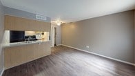 This single-floor unit offers a practical and well-organized layout that makes efficient use of its space. The living area is open and inviting, featuring wood-look laminate flooring, neutral taupe-toned walls, and a textured ceiling with a ceiling fan that promotes airflow and comfort. A large window with vertical blinds allows natural light to fill the room, creating a bright and airy atmosphere. Adjacent to the living space, the kitchen is accessible via a pass-through counter with a wood-paneled partition, adding a warm, functional divider between the two areas. The kitchen is equipped with light wood cabinetry, a double-basin stainless steel sink, a gray laminate countertop, and black appliances including a refrigerator, electric range, and dishwasher. Overhead fluorescent lighting ensures the kitchen is well-lit for cooking and meal preparation. The bathroom features a clean, all-white tub and shower surround with a built-in shelf niche, chrome fixtures, and a standard toilet, offering a straightforward and functional design. Wood-look flooring extends consistently throughout the unit, tying the spaces together with a cohesive aesthetic. The overall design is clean and functional, ideal for everyday living.