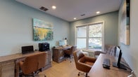 This single-floor office space is thoughtfully designed to accommodate multiple users simultaneously, offering a functional yet aesthetically refined work environment. The walls are painted in a calming sage-green tone that lends the room a relaxed, focused atmosphere, complemented by recessed lighting that ensures even illumination throughout.

Two rustic wood desks with louvered panel detailing serve as the primary workstations, each equipped with desktop computers and additional peripherals, including a multifunction printer. Brown leather rolling office chairs accompany each desk, adding a warm, professional touch. A third wood desk and additional seating are positioned to further expand the room's working capacity.

A large window allows abundant natural light to filter in, offering an outdoor view of lush greenery and a landscaped courtyard. A tufted gray accent chair and a patterned side stool near the window create a comfortable secondary seating area. A woven area rug anchors the space and adds textural warmth underfoot.

Decorative elements include a vibrant large-format landscape painting displayed prominently on one wall, and a trio of wall-mounted geometric planters holding greenery on another. A low wood credenza provides additional storage. Together, these features create a well-balanced, inviting office environment.