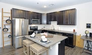 This well-appointed kitchen showcases a cohesive design centered around rich, dark espresso-toned cabinetry that extends from floor to ceiling, providing generous storage throughout the space. The upper and lower cabinets are complemented by light gray countertops, creating a striking contrast that gives the kitchen a contemporary, polished look. A central island serves as both a functional workspace and a casual dining area, accommodating two woven bar stools and offering additional counter space for meal preparation. Stainless steel appliances — including a French door refrigerator, a range with an over-the-range microwave, and a dishwasher — contribute a sleek, modern aesthetic. A dedicated sink area along the back wall features a clean subway tile backsplash, adding texture without overwhelming the design. The light wood-look flooring ties the space together, offering warmth against the darker cabinetry. Recessed lighting provides functional illumination throughout, ensuring a bright and inviting atmosphere. The overall layout is thoughtfully organized for both everyday cooking and entertaining.
