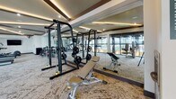 This fitness center is a thoughtfully designed, single-level facility that offers a complete workout experience within a modern and upscale environment. The space is defined by its striking architectural details, including a coffered ceiling with warm recessed accent lighting and bold dark wood beams that lend a refined, contemporary character throughout.

One of the facility's most notable features is its expansive floor-to-ceiling glass wall system with dark-framed panels, which floods the interior with abundant natural light and provides open views of the surrounding outdoor greenery and adjacent buildings. The patterned, low-pile carpet flooring adds texture and comfort underfoot while maintaining a clean, polished aesthetic.

The fitness center is comprehensively outfitted to support a wide variety of training goals. The strength training area includes a cable and power rack machine with free weights and adjustable benches, as well as multiple selectorized resistance machines designed for targeted muscle training. The cardio section features a row of treadmills positioned along the glass wall, offering an engaging view during workouts. Additional equipment includes stability balls and other functional training accessories.

Full-length mirrors line one wall, enhancing the sense of spaciousness and allowing users to monitor their form. A wall-mounted television is provided for entertainment. Entry to the facility is through sleek glass doors, with posted fitness center rules ensuring a structured and respectful environment for all users.