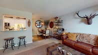 This single-floor residence showcases a cohesive rustic-eclectic aesthetic throughout its thoughtfully arranged layout. The entry hallway features wood-look flooring and conveniently houses a stacked washer and dryer unit, maximizing functionality from the moment you step inside.

The kitchen is outfitted with white shaker cabinetry, granite countertops, stainless steel appliances including a range, microwave, and dishwasher, and a double sink. A peninsula provides additional counter space and casual seating potential, while wood-look flooring ties the space together.

Adjacent to the kitchen, the dining area features warm hardwood-style flooring, a trestle dining table with upholstered chairs, globe pendant lighting, and eclectic wall décor including a round mirror and oversized clock. An open wire shelving unit serves as a stylish room divider between dining and living areas.

The living room offers carpet flooring, a large window with outdoor greenery views, and distinctive décor including a mounted stag head and a leather sofa. Two bedrooms continue the rustic theme—one with a four-poster bed and the other with a large window admitting ample natural light. A dedicated desk nook with an adjacent full bathroom completes the layout.