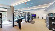 This expansive fitness center offers a generously sized, single-floor layout designed to accommodate a comprehensive range of workout disciplines. The space features a contemporary aesthetic, with soft blue accent ceilings, recessed linear lighting, and concrete-finished columns that lend an upscale, modern character throughout.

The cardio zone is lined with multiple treadmills positioned along a floor-to-ceiling window wall, allowing natural light to flood the space while providing outdoor views during workouts. Adjacent to this area, a dedicated strength training section houses multi-station cable and power rack systems — including Octagon-branded equipment — along with a full dumbbell rack, adjustable benches, and weighted medicine balls organized on wall-mounted shelving. Large mirrors line the walls, enhancing the sense of openness and aiding in form correction.

A separate area accommodates stationary cycling bikes with integrated screens, along with additional cable machines and selectorized strength equipment. The flooring transitions between neutral carpeting and protective rubber mats in high-use zones, offering both comfort and safety. A mounted flat-screen television, water fountain, and staircase access round out the amenities. The overall ambiance is clean, well-lit, and professionally appointed, reflecting a facility built for serious fitness use.
