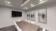 This contemporary conference room delivers a polished and professional environment designed to support productive meetings and collaboration. At its center sits a generously sized rectangular table finished in a warm wood-tone laminate, paired with white ergonomic chairs that offer both comfort and a clean aesthetic. The flooring consists of multi-toned modular carpet tiles in shades of grey, blue, and purple, adding subtle texture and visual depth to the space.

The ceiling features a recessed tray design, fitted with a combination of linear LED strip lighting and recessed can lights, which together provide balanced, glare-free illumination throughout the room. A large wall-mounted flat-screen display serves as the primary presentation focal point, while built-in cable management ports integrated into the tabletop ensure a tidy and functional setup.

A curated set of large-scale abstract artworks in muted grey and white tones adorns the walls, lending an elevated, gallery-like quality to the room. Floor-to-ceiling glass partition walls with dark metal framing connect the space to adjacent meeting rooms and common areas, promoting visual openness and transparency while maintaining acoustic separation. The overall design palette of soft whites, warm neutrals, and dark accents creates a refined, focused atmosphere suited to professional use.