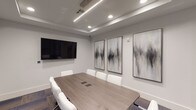 This contemporary conference room delivers a polished and professional environment designed to support productive meetings and collaboration. At its center sits a generously sized rectangular table finished in a warm wood-tone laminate, paired with white ergonomic chairs that offer both comfort and a clean aesthetic. The flooring consists of multi-toned modular carpet tiles in shades of grey, blue, and purple, adding subtle texture and visual depth to the space.

The ceiling features a recessed tray design, fitted with a combination of linear LED strip lighting and recessed can lights, which together provide balanced, glare-free illumination throughout the room. A large wall-mounted flat-screen display serves as the primary presentation focal point, while built-in cable management ports integrated into the tabletop ensure a tidy and functional setup.

A curated set of large-scale abstract artworks in muted grey and white tones adorns the walls, lending an elevated, gallery-like quality to the room. Floor-to-ceiling glass partition walls with dark metal framing connect the space to adjacent meeting rooms and common areas, promoting visual openness and transparency while maintaining acoustic separation. The overall design palette of soft whites, warm neutrals, and dark accents creates a refined, focused atmosphere suited to professional use.