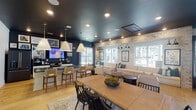 This well-appointed single-floor community amenity space combines modern design with a warm, inviting atmosphere throughout its interconnected zones. The open-concept main area seamlessly integrates a dining space, lounge, and kitchen, all unified by striking deep navy ceilings that contrast beautifully with light hardwood flooring. The dining area features a substantial wooden table with black spindle chairs, anchored by a patterned area rug, while the lounge is furnished with generous cream sofas accented with navy and teal throw pillows. Exposed brick detailing runs along one wall of the lounge, adding warmth and texture to the space. Large picture windows along the lounge walls offer direct views of the outdoor pool and surrounding courtyard, flooding the interior with natural light. The kitchen is thoughtfully designed with white shaker cabinetry, dark stone countertops, gold hardware, a farmhouse sink, and stainless steel appliances — including a French door refrigerator and professional-grade range — all illuminated by oversized conical pendant lights. A dedicated business center features long work surfaces equipped with desktop computers, counter-height bar seating along window ledges, and French doors leading to the exterior, bathed in deep teal tones for a focused yet stylish work environment.