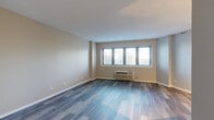 Located at 1101 Iroquois Ave in Naperville, IL, this single-floor unit offers a well-designed, move-in-ready layout with a clean, contemporary aesthetic throughout. The living area is generously proportioned, finished with stylish wood-look luxury vinyl plank flooring in a cool gray tone that extends seamlessly throughout the space. A wide multi-pane window with blinds allows abundant natural light to fill the room while offering views of the surrounding greenery, and a wall-mounted HVAC unit ensures year-round comfort. The open layout connects the living space to a compact, efficiently arranged kitchen equipped with granite-style countertops, white cabinetry, a stainless steel dishwasher, and a stove, all accented by track lighting overhead. A hallway with a sliding barn-style door leads to additional areas of the unit, maintaining a sense of separation while keeping the overall floor plan open and connected. The bathroom is neatly appointed with a white vanity featuring granite-style countertops, brushed nickel hardware, a tiled shower-tub combination, and matching vinyl plank flooring. Neutral wall tones in warm beige and soft gray unify the space, creating a calm and inviting atmosphere that is both functional and visually cohesive.