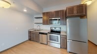 Located at 511 3rd Ave SW in Rochester, Minnesota, this single-floor unit offers a clean, modern layout designed for comfortable everyday living. The open living area is finished with light wood-tone flooring and neutral-toned walls, creating a bright and welcoming atmosphere. A large window dressed with vertical blinds allows natural light to fill the space, while a sliding door provides additional access and openness to the room.

The kitchen is well-appointed with rich, dark wood cabinetry, granite-style countertops, and a full suite of stainless steel appliances including a refrigerator, electric range, built-in microwave, and dishwasher. Open shelving adds a contemporary touch and provides additional storage options alongside the ample cabinetry.

The bathroom features a tub and shower combination with a curtain rod, a toilet, and a vanity with a granite countertop and dark wood cabinetry. Light tile flooring and neutral gray walls give the bathroom a clean, functional feel. An overhead ventilation unit and a well-positioned mirror round out the space.

Additional areas visible in the layout include a carpeted room that functions as a bedroom or secondary living space, along with closet access. The unit's efficient floor plan maximizes usability while maintaining a cohesive, contemporary aesthetic throughout.