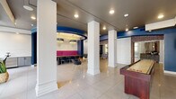 Located at 305 Axis Dr in Louisville, KY, this single-floor community amenity space is designed to serve a variety of social and functional needs through several well-defined zones unified by a cohesive contemporary aesthetic. Polished large-format tile flooring and crisp white structural columns anchor the open layout, while navy blue accents, exposed brick archways, and dark ceilings create visual contrast throughout.

The lounge area features a built-in entertainment wall with a flat-screen TV set against a bold blue accent backdrop, flanked by illuminated shelving with herringbone-patterned tile inserts. Gray upholstered seating, a round wood coffee table, and a decorative chandelier lend a refined yet relaxed atmosphere, with glass doors offering views to an outdoor courtyard.

A distinctive dining nook is defined by a curved tray ceiling with a navy band and two statement crystal chandeliers. Bold magenta banquette seating and a communal wood table create an intimate gathering spot beside a kitchenette and bar-height seating near large windows.

The gaming area includes a full shuffleboard table, wall-mounted TVs in blue frames, and an arcade space accessible through an exposed brick archway. A separate business center offers carpeted flooring, a workstation with computer, wall-mounted TV, and additional seating for focused work.