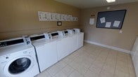 This shared laundry facility offers a practical and well-organized space designed to serve multiple users efficiently. The room features a row of top-loading washing machines and dryers lined neatly along the wall, providing ample capacity for communal use. The light-colored tile flooring is easy to maintain and contributes to the room's clean, utilitarian aesthetic. The neutral beige walls keep the atmosphere simple and bright. A series of informational instruction sheets are mounted along the back wall above the machines, offering clear guidance on proper machine usage. A bulletin board on the adjacent wall serves as a community communication hub, displaying posted notices and announcements. A paper towel or document dispenser is also mounted nearby for added convenience. The space is single-story and thoughtfully laid out to allow users to move comfortably between machines. Overall, the facility is functional, tidy, and equipped with everything needed for a smooth and straightforward laundry experience.