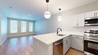 This well-designed 1-bedroom, 1-bathroom unit offers a thoughtful layout across a single floor, combining modern aesthetics with practical functionality. The open-concept living and kitchen area forms the heart of the unit, finished with warm hardwood-style flooring that extends throughout the main living spaces. The kitchen is a standout feature, equipped with stainless steel appliances including a range, microwave, dishwasher, and refrigerator. Two-tone cabinetry pairs white upper cabinets with grey lower cabinets, complemented by crisp white countertops and a peninsula island with a built-in sink. Stylish globe pendant lights add a contemporary touch overhead. The living area benefits from multiple windows that invite natural light and connect seamlessly to the kitchen. The spacious primary bedroom features soft carpeting, generous wall space, and a window that brings in additional natural light. A dedicated closet provides convenient storage. The bathroom is finished with wood-style flooring, a full tub-shower combination, and matte black fixtures that lend a modern, cohesive look. Neutral grey-toned walls throughout the unit create a clean, versatile backdrop suited to a variety of interior styles.