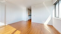 Located at 1190 Mission Street in San Francisco's SoMa neighborhood, this single-floor residence offers a clean, modern aesthetic defined by crisp white walls and warm hardwood flooring that flows seamlessly throughout the living areas. The open layout connects the main living space to a functional galley-style kitchen, which is outfitted with white cabinetry, dark countertops, a stainless steel sink, and white appliances including a refrigerator and stove. Natural light plays a prominent role in the living and bedroom areas, where large windows frame sweeping views of the San Francisco skyline, filling the rooms with abundant sunlight throughout the day. A vertical panel radiator provides efficient heating along one wall of the bedroom, complementing the airy, sunlit atmosphere. The bathroom is finished in all-white tile, featuring a full soaking tub with an overhead shower, built-in open shelving for storage, a toilet, and a mirrored medicine cabinet — all contributing to a clean, minimalist feel. The overall design of the space emphasizes simplicity and functionality, with a neutral palette that allows the natural light and city views to take center stage. This well-appointed residence offers a comfortable urban living experience in one of San Francisco's most vibrant and centrally located neighborhoods.