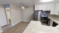 This single-floor space offers a clean, modern aesthetic characterized by neutral tones, wood-look plank flooring, and textured ceilings throughout. The open-concept kitchen is equipped with a full suite of stainless steel appliances, including a refrigerator, electric range, and built-in microwave. White shaker-style cabinetry provides ample storage, while light granite-style countertops offer generous prep space. A breakfast bar or counter peninsula adds both functionality and a casual dining option. The adjoining living and dining area is bright and open, featuring a ceiling fan with integrated lighting that enhances both comfort and ambiance. Neutral gray walls and crisp white baseboards create a cohesive, contemporary look that complements the warm tones of the flooring. The bedroom continues the same refined palette, with a ceiling fan, a closet with white bi-fold doors, and direct access to what appears to be a well-lit bathroom. The consistent use of updated materials and finishes throughout the space reflects a thoughtfully refreshed interior, making it an inviting and functional living environment.