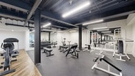 Located on Wilshire Boulevard in Los Angeles, this fitness facility offers a thoughtfully designed exercise space spread across a single level. The interior is divided into two distinct functional zones, each with its own flooring and aesthetic character. The strength training area features durable speckled rubber flooring and houses a comprehensive array of free weights, including a full dumbbell rack, adjustable benches, a cable machine, and various resistance stations. Large wall mirrors amplify the sense of space and support proper form during workouts. The cardio zone transitions to a warm herringbone-patterned hardwood floor, lending a refined contrast to the industrial ceiling above. This area is equipped with upright bikes, elliptical machines, and treadmills, alongside a wall-mounted display screen. Throughout the facility, the exposed black ceiling with structural steel columns and linear LED lighting creates a modern industrial ambiance. Windows allow natural light to filter in, connecting the space to the surrounding environment. The overall design balances functionality with a sleek, contemporary aesthetic, making it a well-rounded fitness destination.