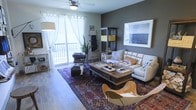 This single-floor living space showcases a thoughtfully designed interior that blends modern and rustic aesthetics. The main living area is anchored by a cream sectional sofa adorned with textured throw pillows, paired with a substantial reclaimed wood coffee table that serves as a focal point of the room. A vibrant, richly patterned traditional area rug grounds the seating arrangement, adding warmth and depth to the space.

The room features dark gray accent walls that create a striking contrast against the lighter furnishings and sheer white curtains. A large framed artwork hangs prominently on one wall, contributing to the curated feel of the space. A decorative ladder leans against the wall, providing both visual interest and functional storage for throws and accessories.

Natural light flows generously through floor-to-ceiling windows and a sliding glass door that opens onto a balcony, offering views of the surrounding area. A ceiling fan with integrated lighting provides both comfort and ambient illumination. Additional storage is provided by a dark shelving unit displaying decorative boxes and accessories. Hardwood flooring throughout ties the space together, complementing the eclectic mix of furnishings, including a distinctive leather sling chair.