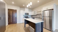 This single-floor residence offers a clean, contemporary design highlighted by light hardwood-style flooring that flows consistently throughout the space. The kitchen serves as the focal point, showcasing white shaker-style cabinetry that extends to the ceiling, complemented by a classic gray subway tile backsplash and dark countertops. A full suite of stainless steel appliances — including a refrigerator, range, microwave, and dishwasher — provides modern functionality. A freestanding center island with a crisp white countertop and dark base offers additional prep space and seating potential, while a trio of pendant lights above adds a stylish, focused glow. The open layout connects the kitchen seamlessly to a generously sized living area, which is bathed in natural light streaming through large French doors topped with a transom window. These doors open directly to a lush green outdoor area, creating an inviting indoor-outdoor connection. The bathroom features a clean vanity with white countertops, shaker-style cabinetry, and a framed mirror, maintaining the home's cohesive, neutral aesthetic. Throughout, recessed lighting and neutral wall tones contribute to a bright, airy atmosphere that is both functional and move-in ready.
