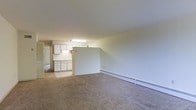 This single-floor residential unit offers a straightforward and functional layout designed to maximize usable living space. The open living area is finished with wall-to-wall carpeting, giving the space a warm and comfortable feel underfoot. A baseboard heating unit runs along the exterior wall, ensuring efficient climate control throughout the colder months. The living area flows naturally toward the kitchen, which is partially separated by a half-wall partition that adds a sense of structure while maintaining an open feel. The kitchen is equipped with white cabinetry both above and below the countertops, providing generous storage space. A sink is positioned beneath the upper cabinets, and overhead fluorescent lighting illuminates the workspace effectively. The neutral white walls throughout the unit create a clean, bright backdrop that allows for easy personalization. A hallway visible from the living area suggests additional rooms beyond, contributing to the overall sense of a practical and well-organized floor plan. The unit's simple, uncluttered design makes it a versatile space suitable for a range of occupants.